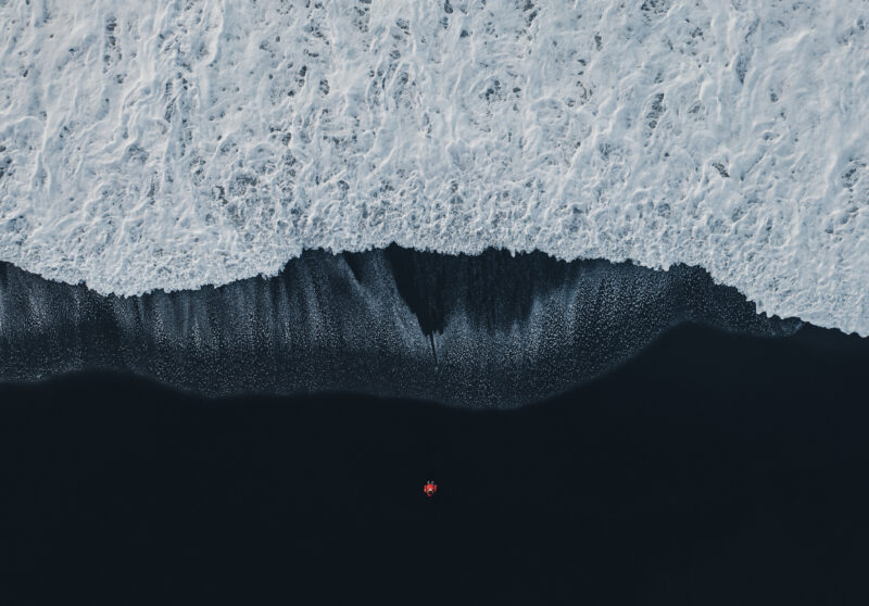 Aerial view of woman in red coat standing on black sand beach in Iceland.